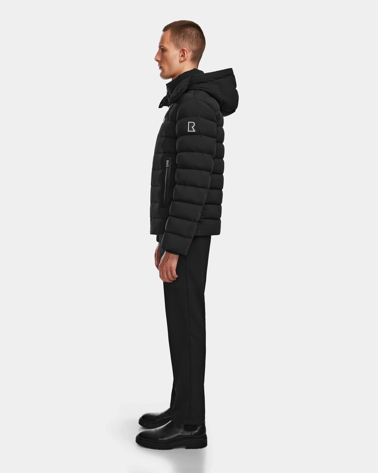 A man stands in profile wearing the Rudsak NAVAN - 6124551 Black quilted down puffer jacket, featuring wind and water-repellent properties, paired with black pants and boots against a plain white background.