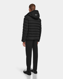 A man faces away wearing the Rudsak NAVAN - 6124551 Black mens quilted down puffer jacket with hood, black pants, and shoes against a plain light background. The jacket features wind- and water-repellent fabric and RDS-certified down fill.