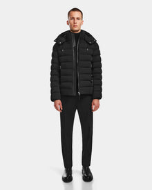 A person stands facing forward wearing the Rudsak NAVAN - 6124551 Black mens quilted down puffer jacket with a hood, paired with black pants, black shoes, and a black shirt against a plain white background.