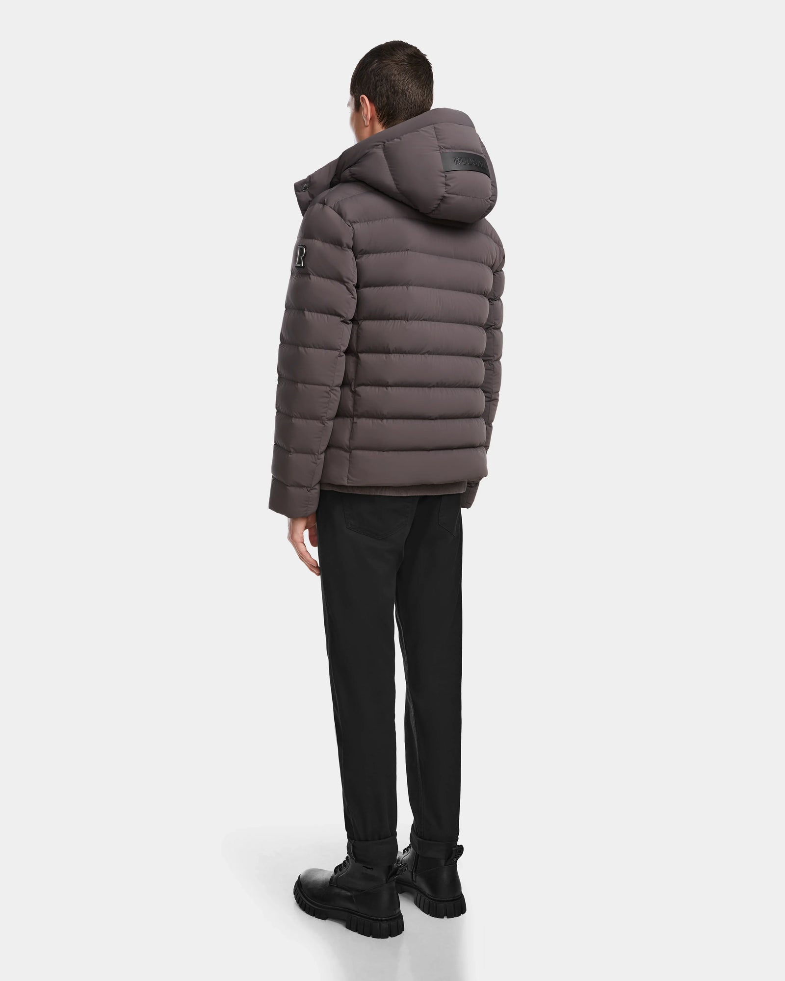 A person stands with their back to the camera, wearing the Rudsak NAVAN - 6124551 Grey mens quilted down puffer jacket with a hood, black pants, and black boots against a plain white background.