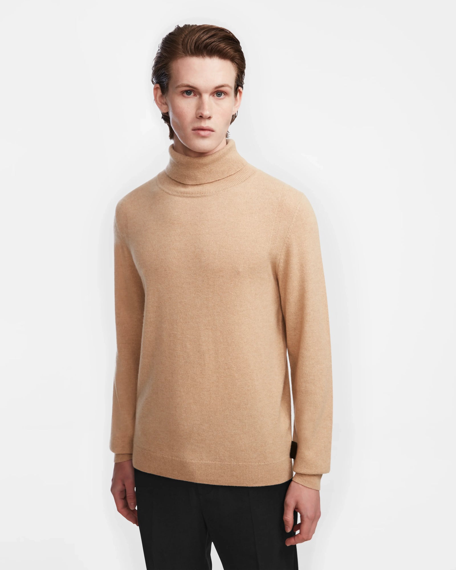 A young man with light skin and brown hair models the Rudsak NICOLAS - 6524528 Camel turtleneck sweater, paired with black pants, standing against a plain white background and facing the camera.