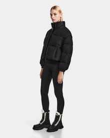 A woman with straight blonde hair and a neutral expression wears the Rudsak OPRAH - 8124708 Black women’s cropped down puffer, paired with black leggings and black fur-trimmed lace-up boots, against a plain white background.