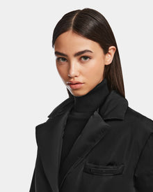 A woman with straight, dark brown hair and light skin models the PAOLA - 8124547 Black weather-resistant blazer by Rudsak over a black turtleneck, looking directly at the camera against a plain light background.