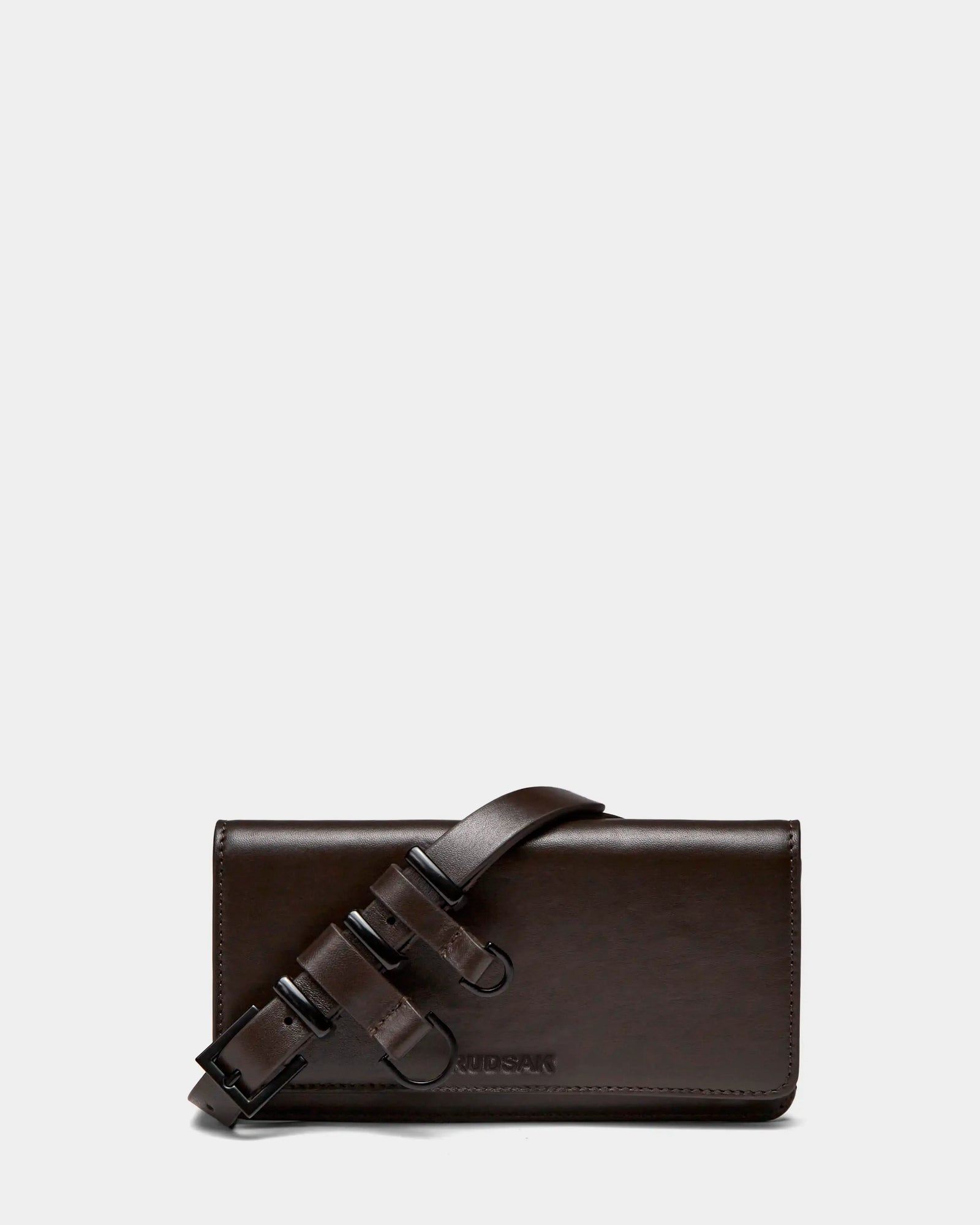 The PREYA - 8324508 Espresso by Rudsak is a rectangular dark brown leather clutch with a wide matching shoulder strap and black metal buckles, displayed on a plain light gray background.