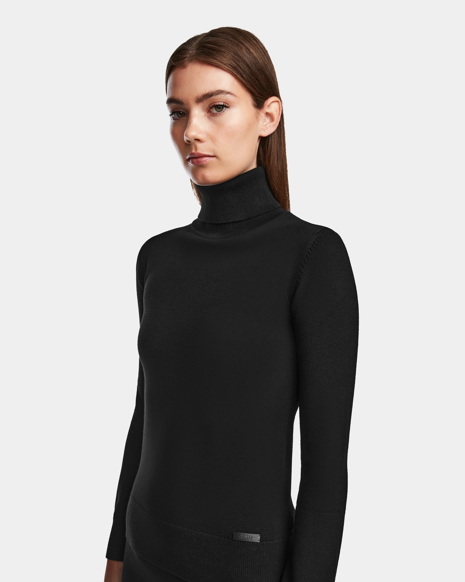 A woman with straight brown hair wears the RALDI - 8524605 Black knit turtleneck by Rudsak, a fitted sweater, standing against a plain white background and looking slightly to the side with a neutral expression.