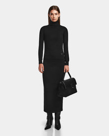A woman faces forward against a white background, wearing the Rudsak RALDI - 8524605 Black womens knit turtleneck sweater with a matching long skirt and black boots, holding a black handbag by her side.