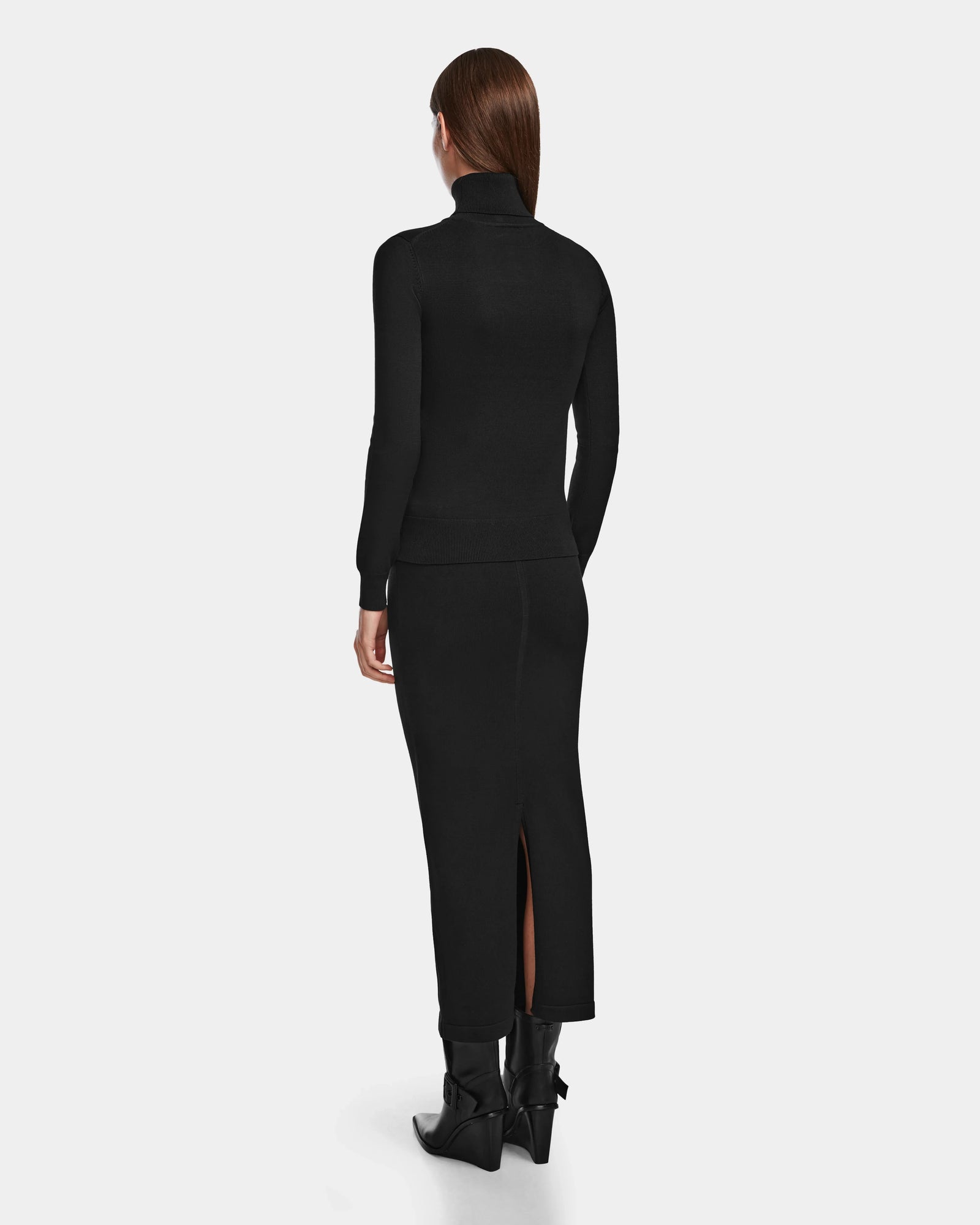 A woman with straight brown hair faces away, wearing the RALDI - 8524605 Black by Rudsak—a fitted metallic thread turtleneck—paired with a long black skirt with a back slit and black heeled boots, against a plain white background.