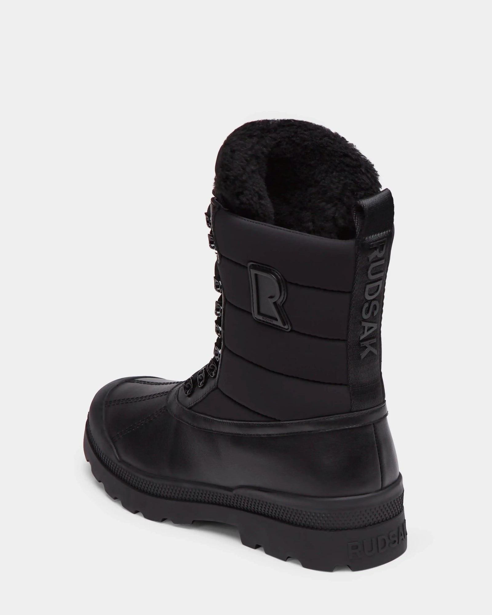 The Rudsak REAGAN - 6224501 Black men’s winter boot features a thick rubber sole, lace-up front, faux fur lining, side branding, and back pull tab. Shown on a plain white background.