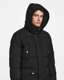 A person wearing the Rudsak RICKY - M123502 Black men's quilted down parka with the hood up, looking slightly to the side against a plain white background.
