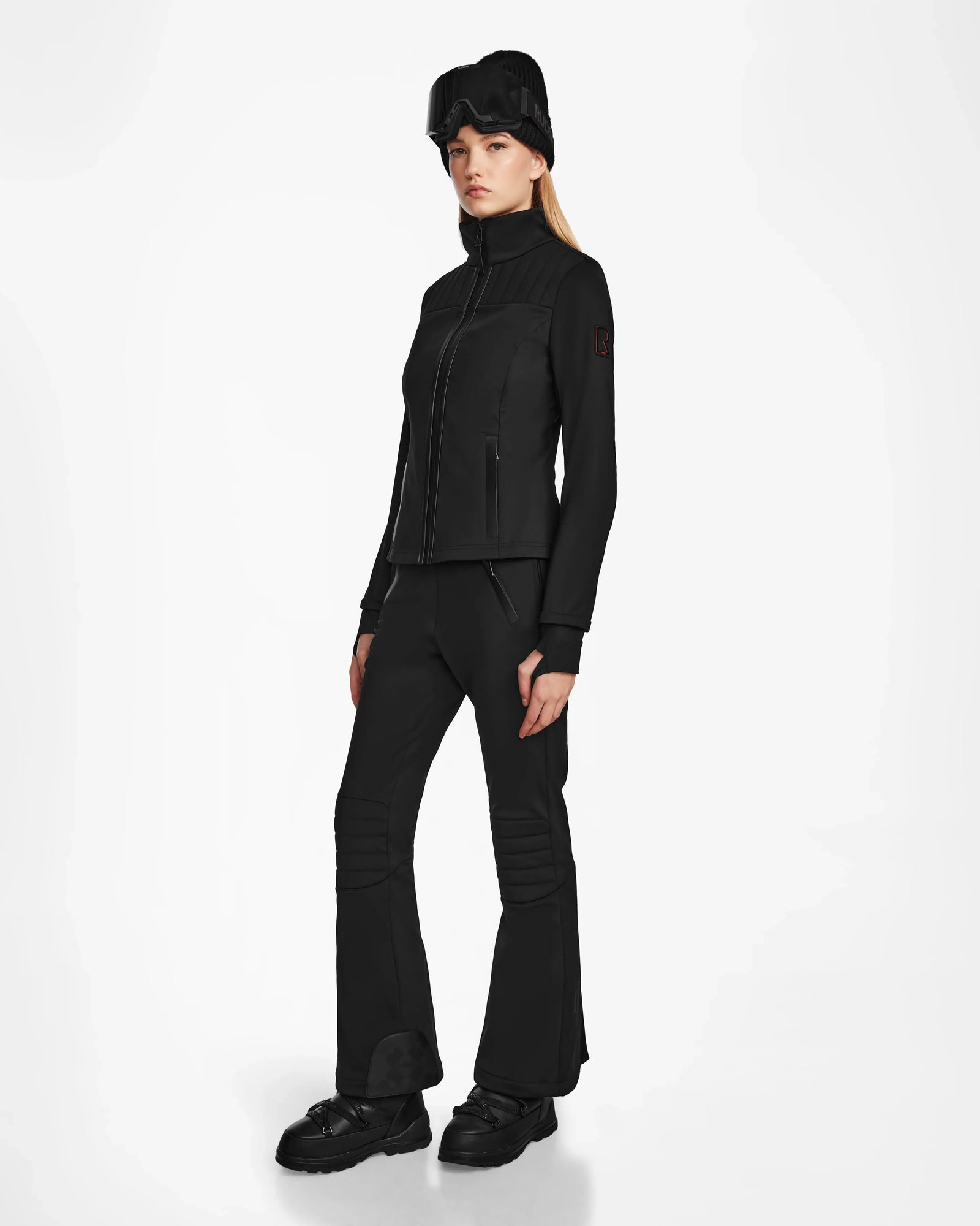 A woman stands against a plain white background wearing the Rudsak ROSALIE - 8124784 Black fitted ski jacket with RECCO rescue technology, paired with flared black pants, black boots, and a black fur hat. She faces slightly to the side, expression neutral.