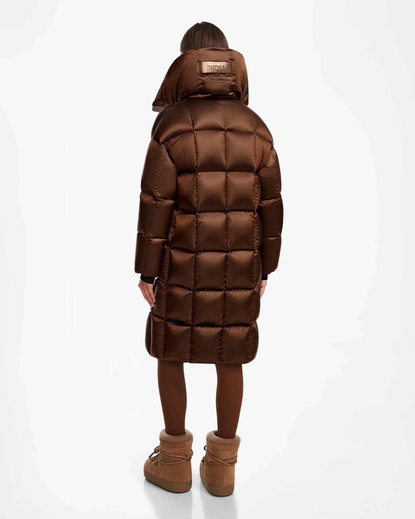 A person stands facing away, wearing the Rudsak SASH R - 8124543 Antique Bronze women’s long down puffer coat with a high collar, dark tights, and brown winter boots against a plain white background.