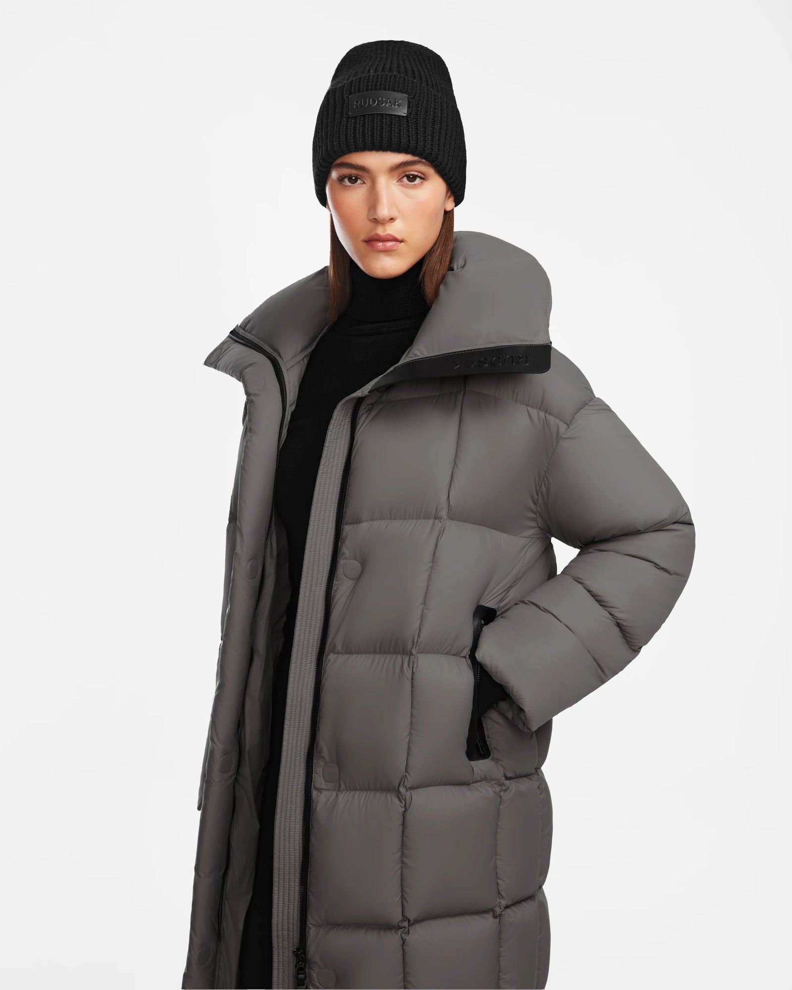 A woman models the Rudsak SASH - 8124536 Light Grey womens long down puffer coat and a black knit beanie, standing against a plain white background and highlighting its stylish winter design with RDS-certified down fill.