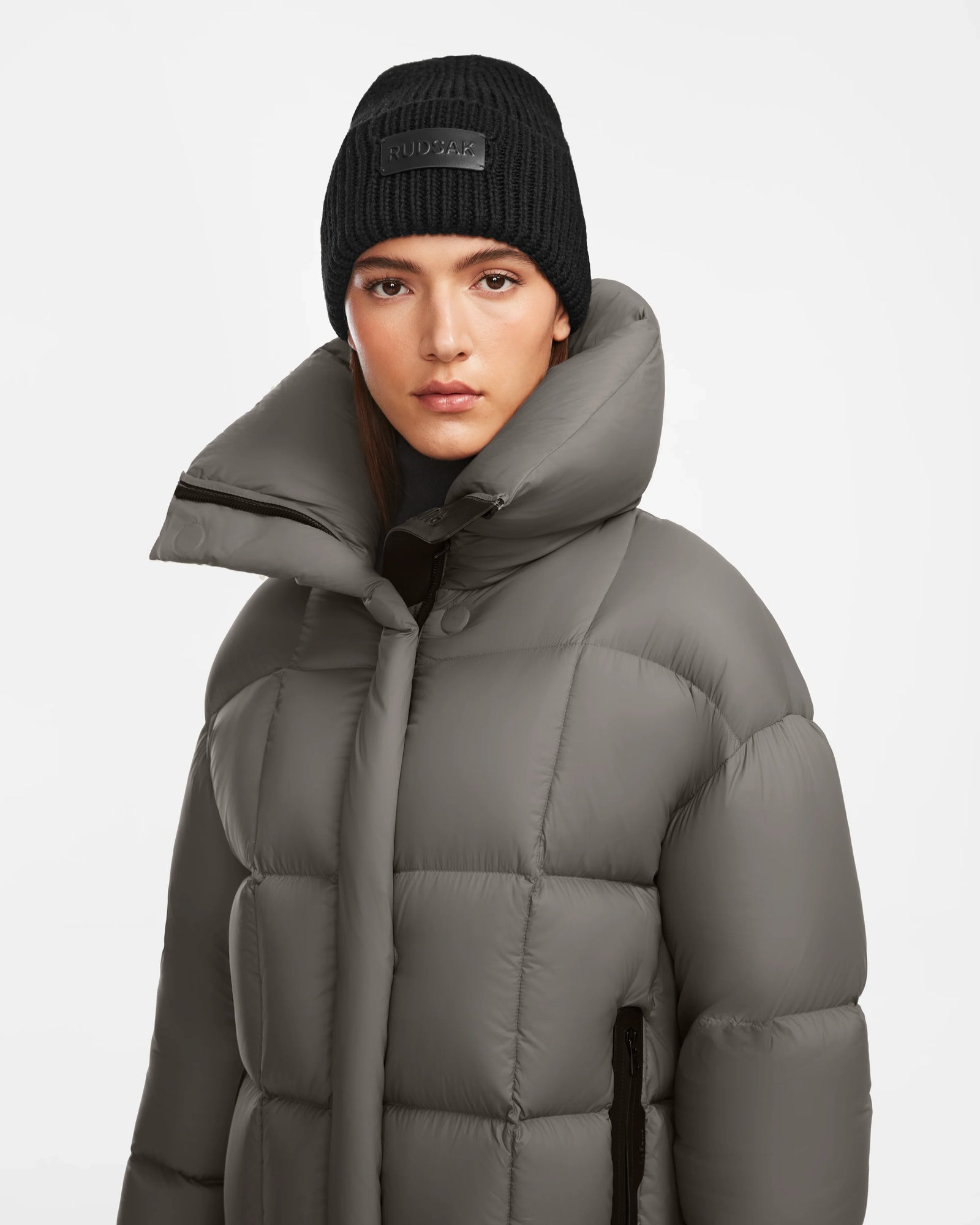 A woman faces the camera with a neutral expression, wearing the Rudsak SASH - 8124536 Light Grey women’s long down puffer coat with a high collar and a black knit beanie, set against a plain white background.