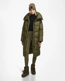 Wearing the Rudsak SASH - 8124536 Loden Green womens long down puffer coat, paired with leggings, a beanie, and boots, a model stands against a plain white background. The coat looks warm and ideal for cold weather.