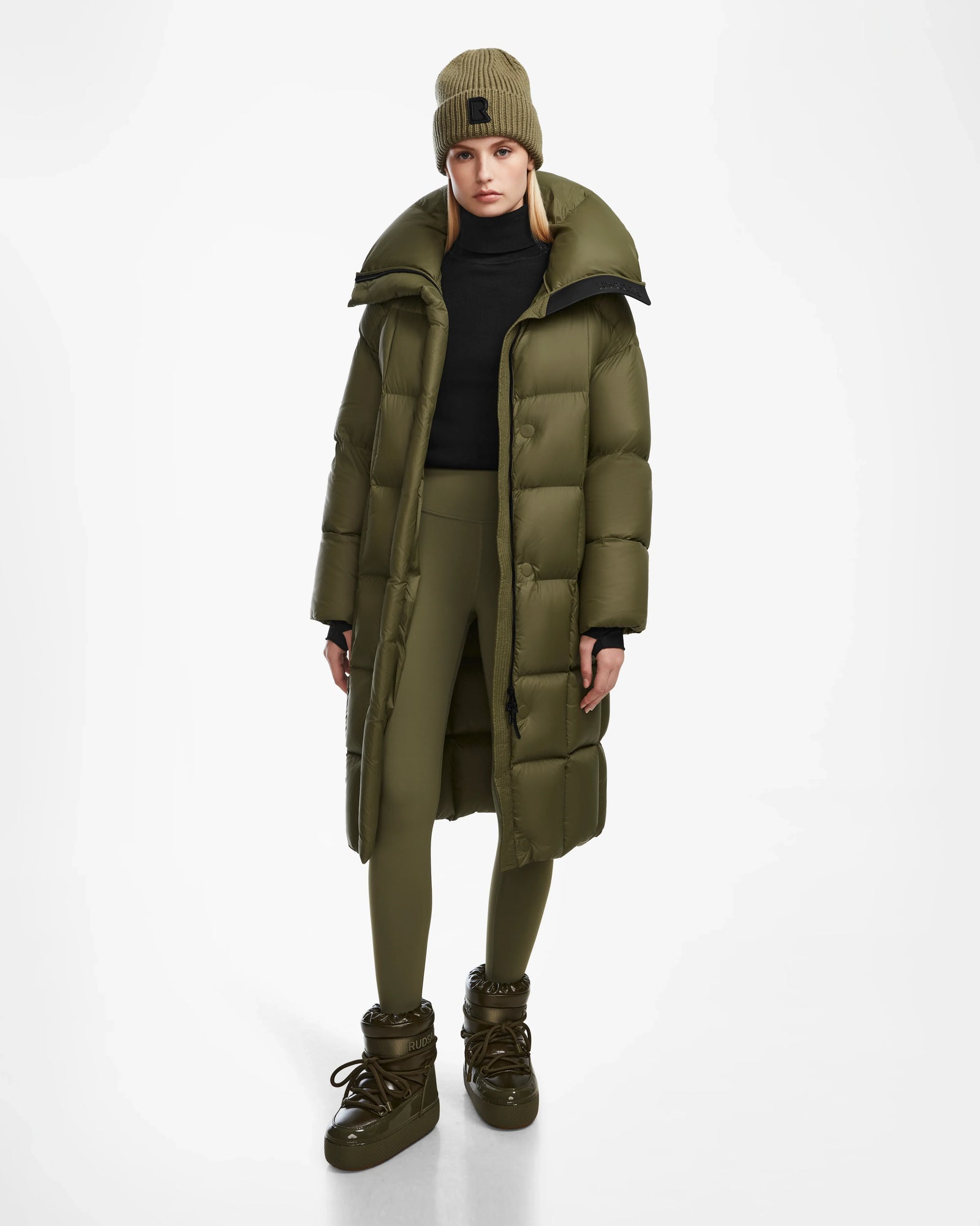 Wearing the Rudsak SASH - 8124536 Loden Green womens long down puffer coat, paired with leggings, a beanie, and boots, a model stands against a plain white background. The coat looks warm and ideal for cold weather.
