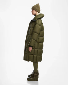 A person stands in profile wearing the Rudsak SASH - 8124536 Loden Green womens long down puffer coat with a matching beanie, leggings, and platform winter boots against a plain white background.