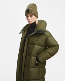 A woman in the Rudsak SASH - 8124536 Loden Green long down puffer coat and a matching knit beanie stands against a white background, looking at the camera with a neutral expression—an ideal winter outerwear look.