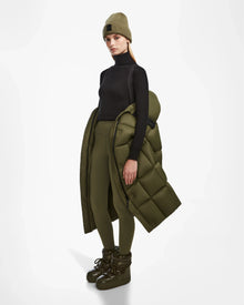 A person stands against a white background wearing Rudsaks SASH - 8124536 Loden Green womens long down puffer coat draped over one arm, paired with matching olive green leggings, a tan beanie, and olive green winter boots for perfect winter style.
