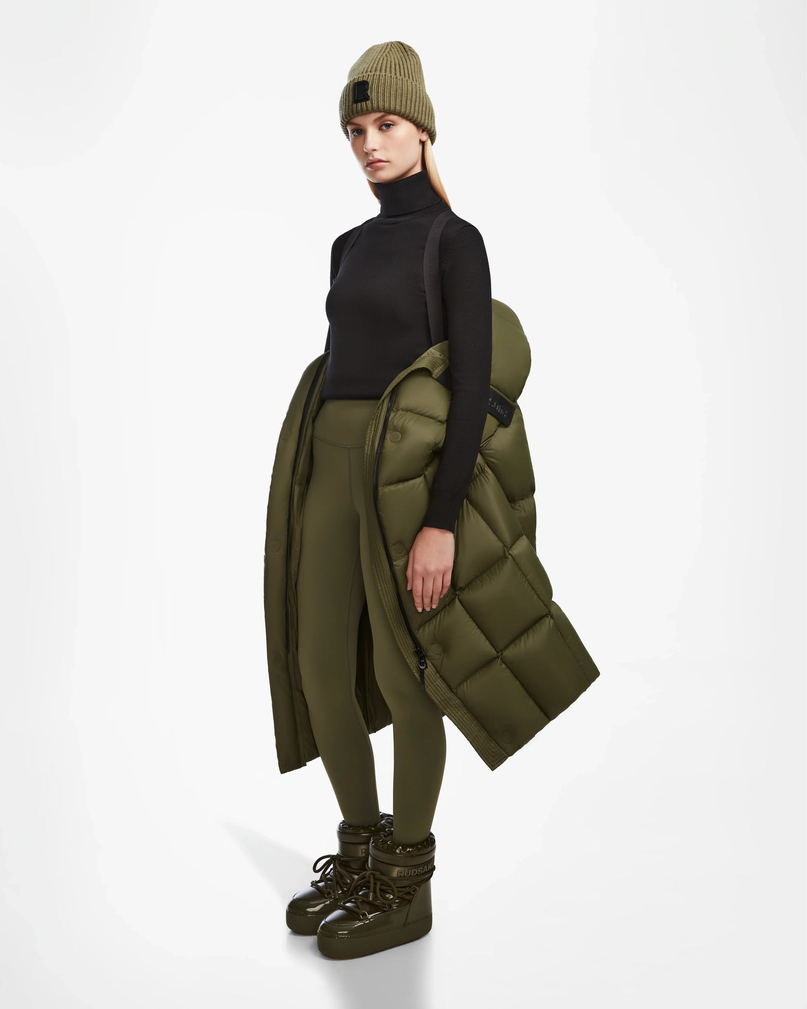 A person stands against a white background wearing Rudsaks SASH - 8124536 Loden Green womens long down puffer coat draped over one arm, paired with matching olive green leggings, a tan beanie, and olive green winter boots for perfect winter style.