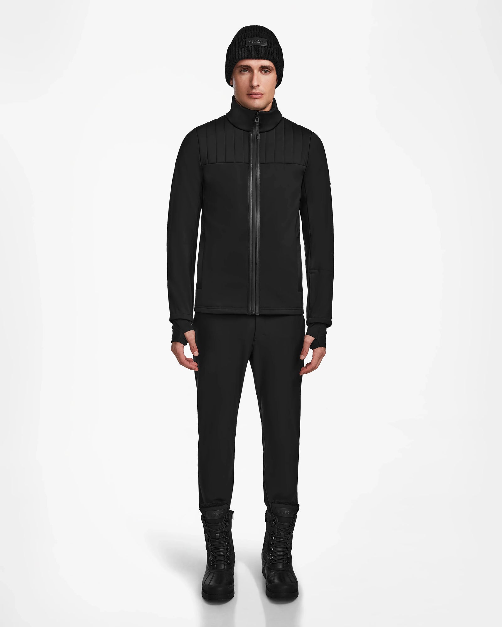 A person stands facing forward, wearing the Rudsak TREVOR - 6124519 Black mens ski jacket, black pants, and black lace-up boots against a plain white background.