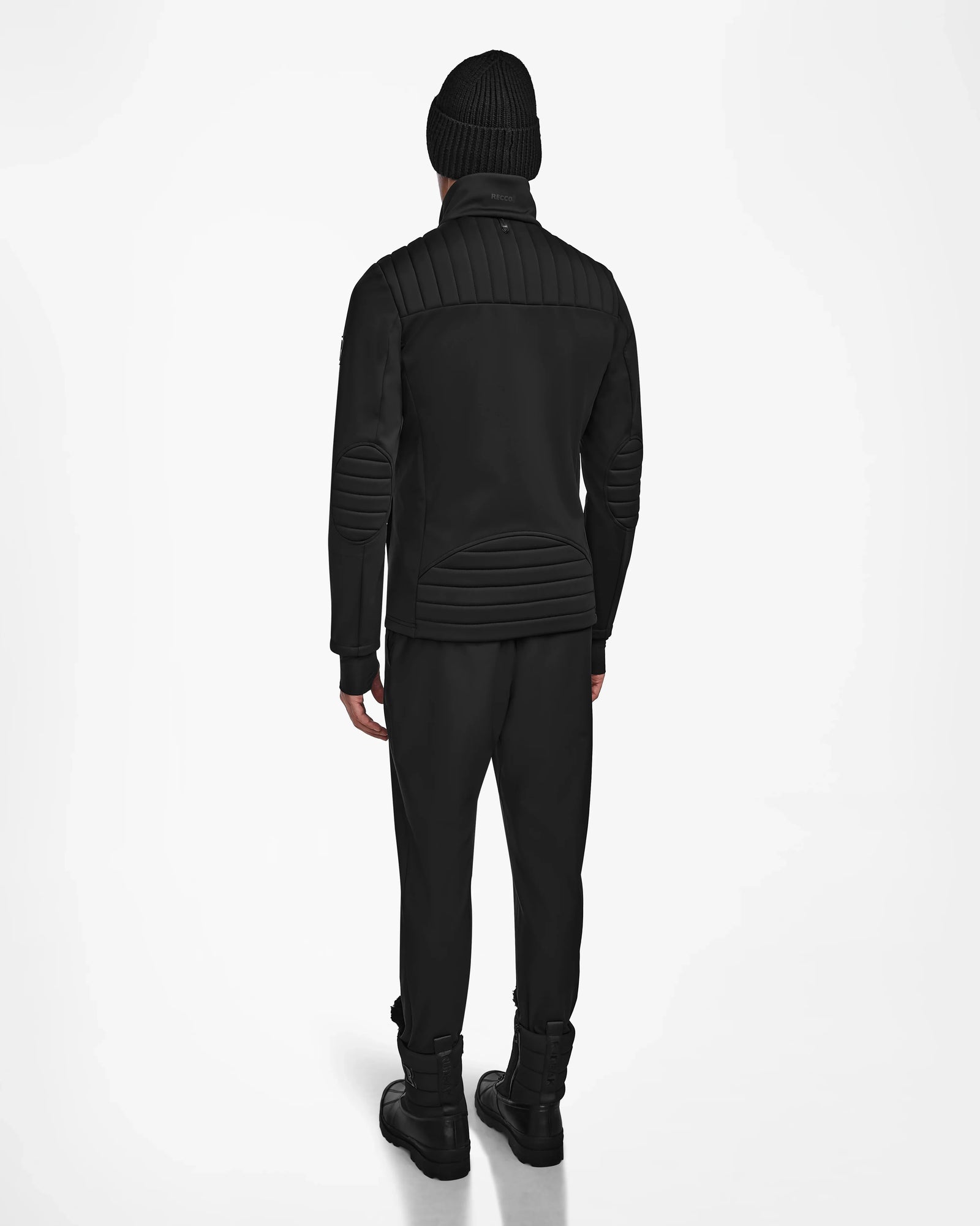 A person in all-black attire, featuring the Rudsak TREVOR - 6124519 Black mens ski jacket, beanie, and boots, stands facing away from the camera against a plain white background.