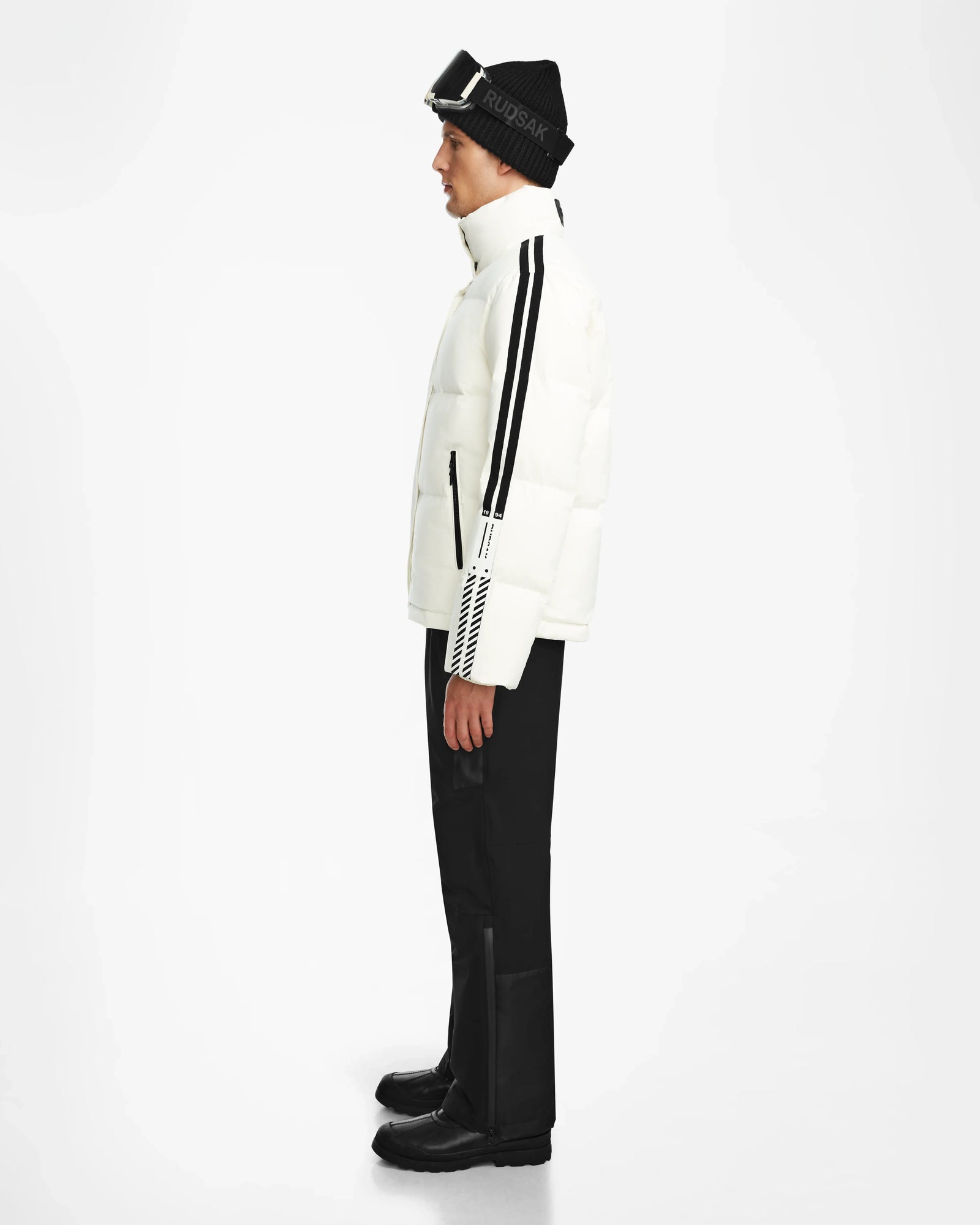 A person in side profile wears the Rudsak USHER - 6124516 Optic White men’s down ski jacket, black pants, black boots, and a black knit hat with goggles on top, standing against a plain white background.
