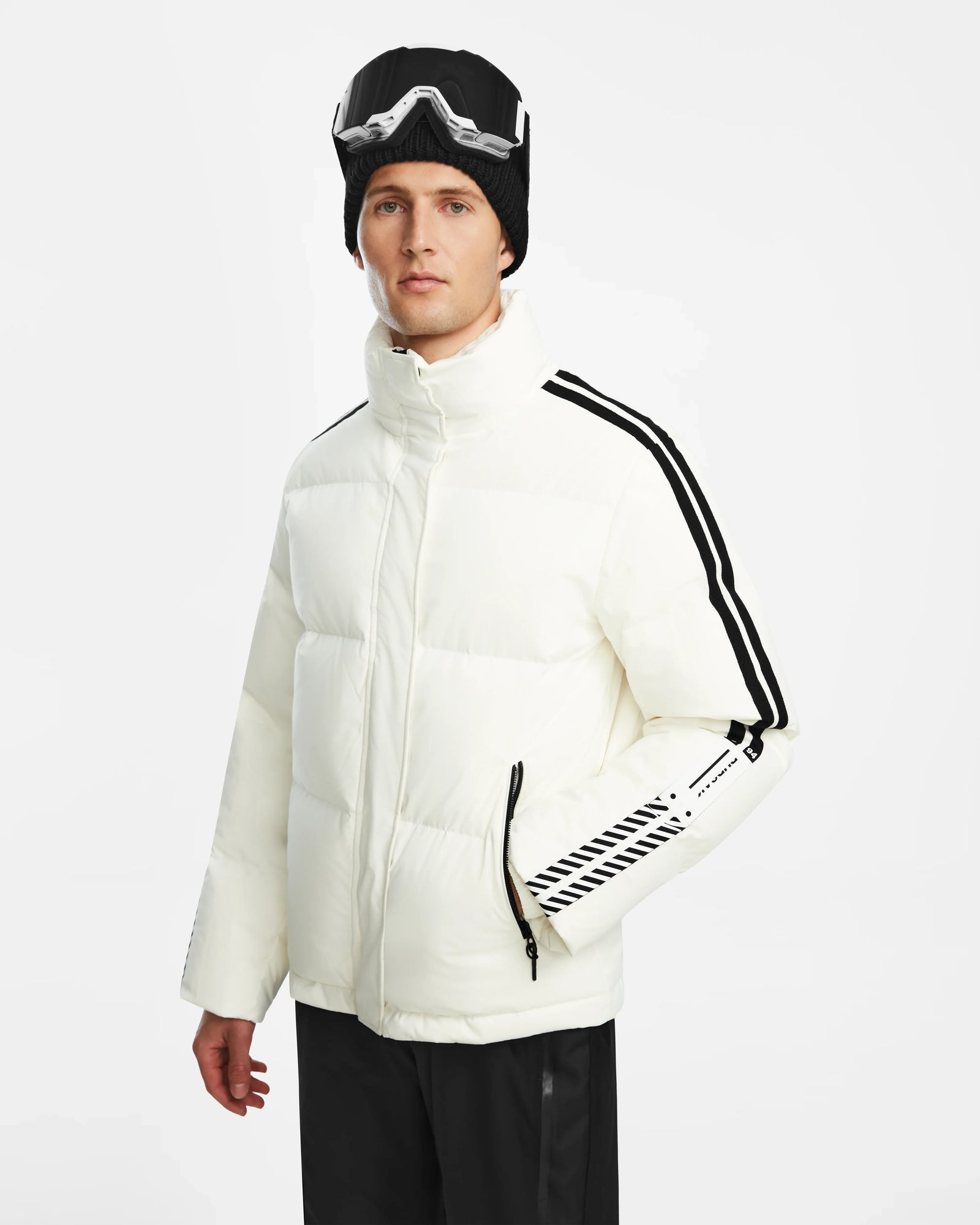 A man wearing the Rudsak USHER - 6124516 Optic White mens down ski jacket with black stripes, black ski goggles on his head, and black pants stands against a plain white background.