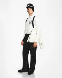Dressed in black ski pants, boots, black goggles, a black hat, and a white long-sleeve top, the person wears Rudsaks USHER - 6124516 Optic White mens down ski jacket with RDS-certified insulation against a white background.