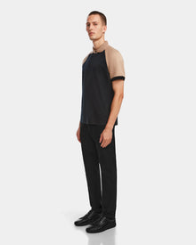 A man stands facing slightly left wearing the Rudsak WALTER - 6524531 Black/Camel premium cotton polo with beige sleeves and a dark body, paired with black pants and sneakers against a plain light background.