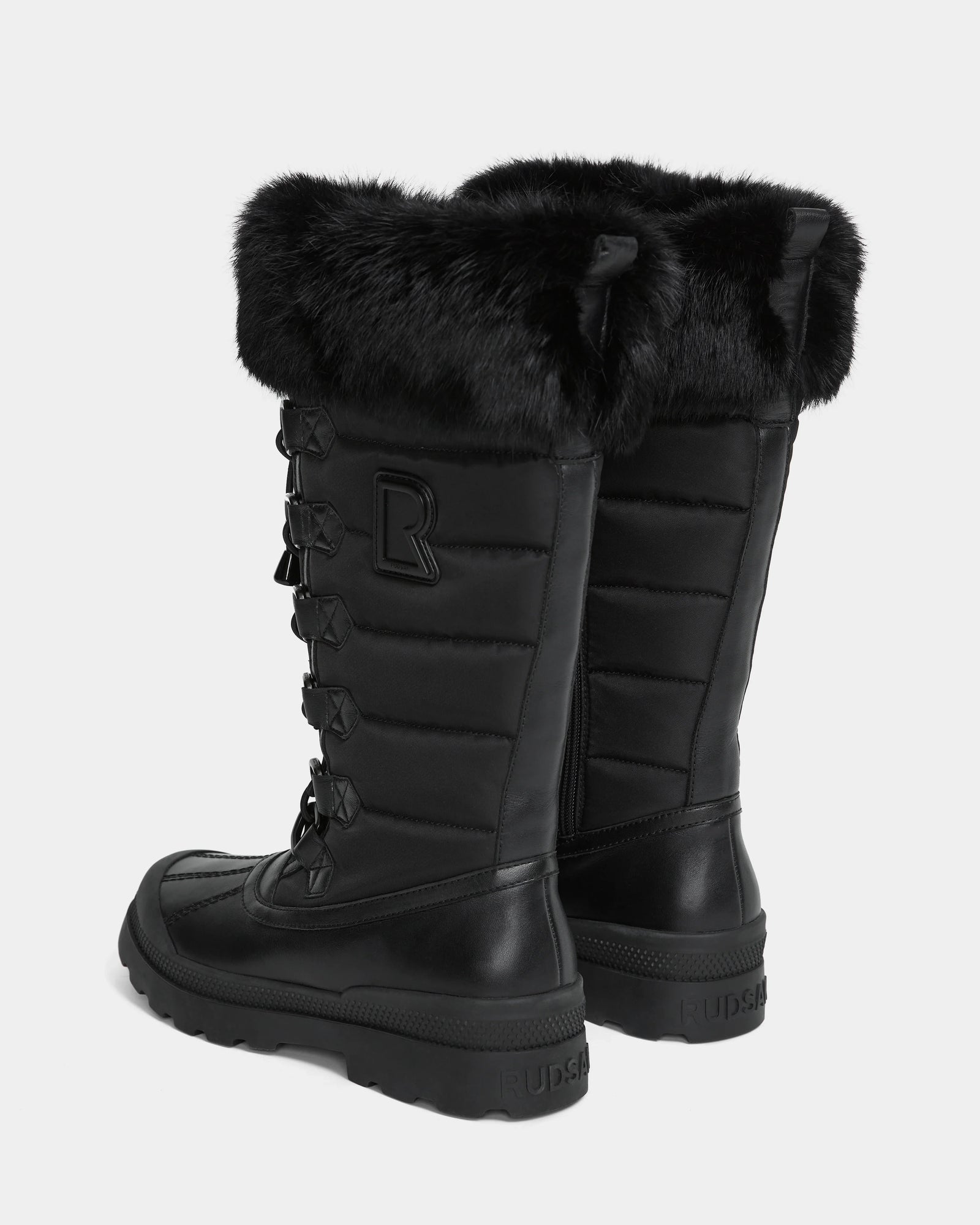 The Rudsak WINDY - L224507 Black is a pair of tall women’s winter boots featuring faux fur trim, quilted sides, thick rubber soles, decorative laces, a side zipper, and an "R" stitched near the top outer side of each boot.