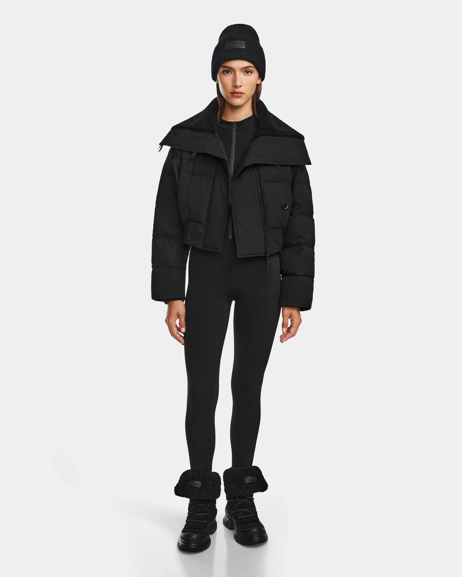 A woman faces forward in a Rudsak YVETTE - 8124710 Black corduroy puffer jacket with a wide collar, paired with a black beanie, leggings, and fur-trimmed boots, highlighting chic winter style against a plain light background.