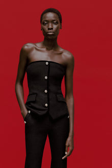 Black strapless bustier top with pearl button front closure and side pockets, worn with matching trousers.