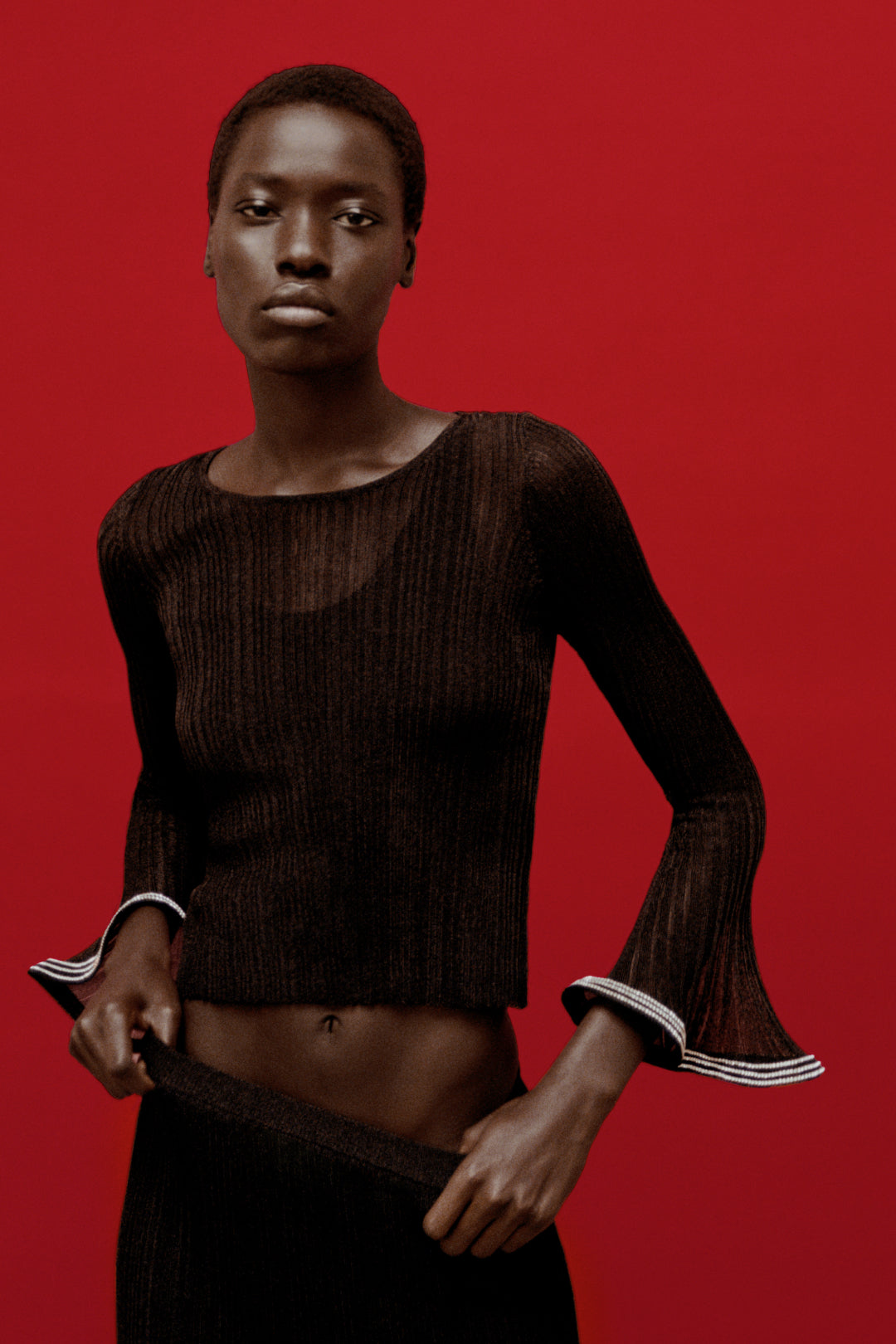 Black metallic plissÃ© top with long flared sleeves and striped trim cuffs on red background.