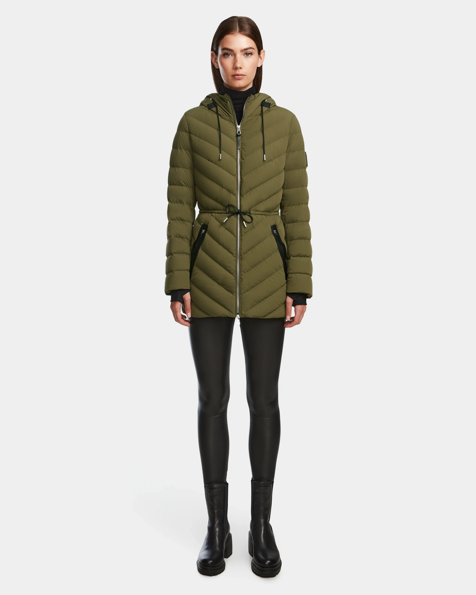 A woman faces forward wearing the Rudsak FRANCINE - 8124722 Loden Green quilted puffer jacket with a hood, drawstring waist, and zip closure, paired with black leggings and ankle boots against a white background.