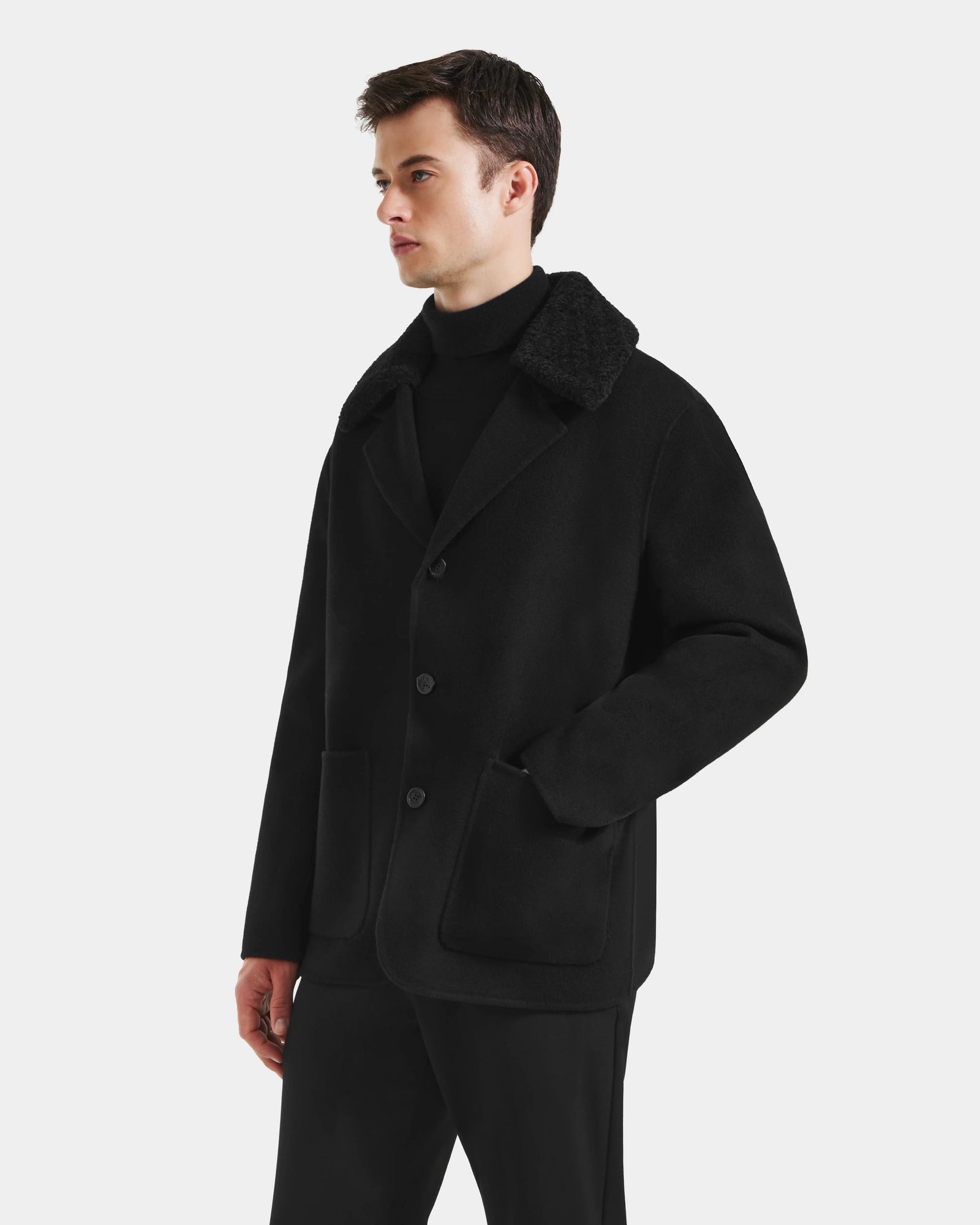 A man wearing the Rudsak AIDEN - 6125558 Black men’s wool-blend coat with large front pockets and a black turtleneck stands against a plain white background.