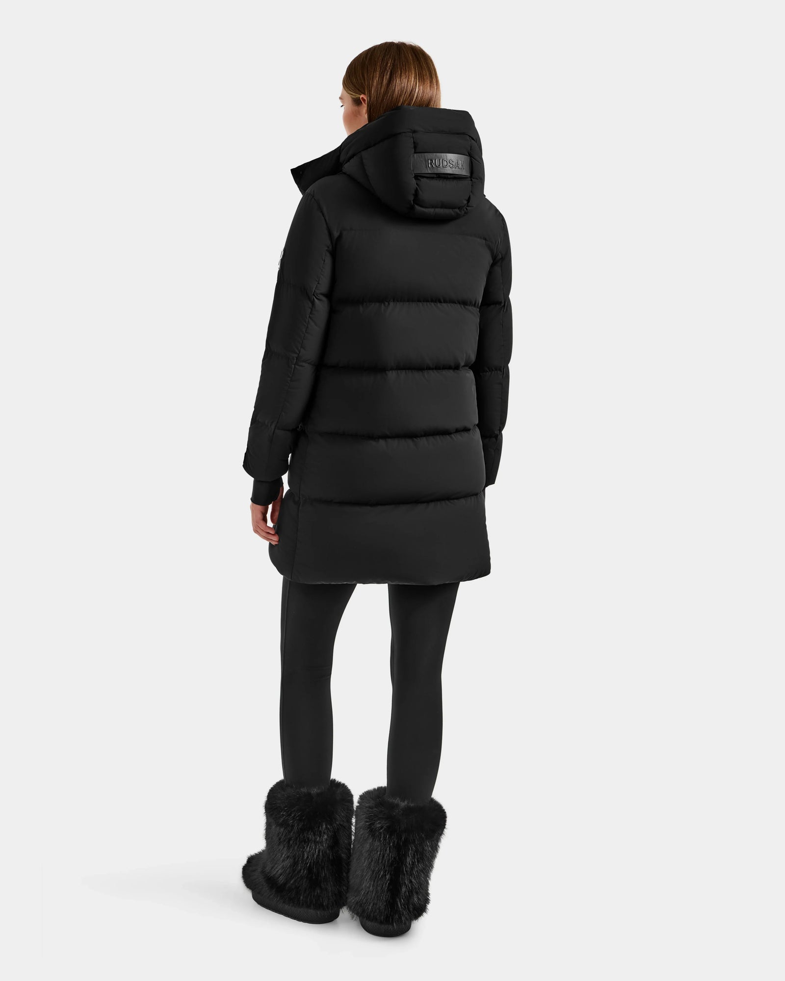 A person wearing the Rudsak ALESSANDRA M - L125552 Black women's down puffer, black leggings, and black furry boots stands with their back to the camera against a plain light background.