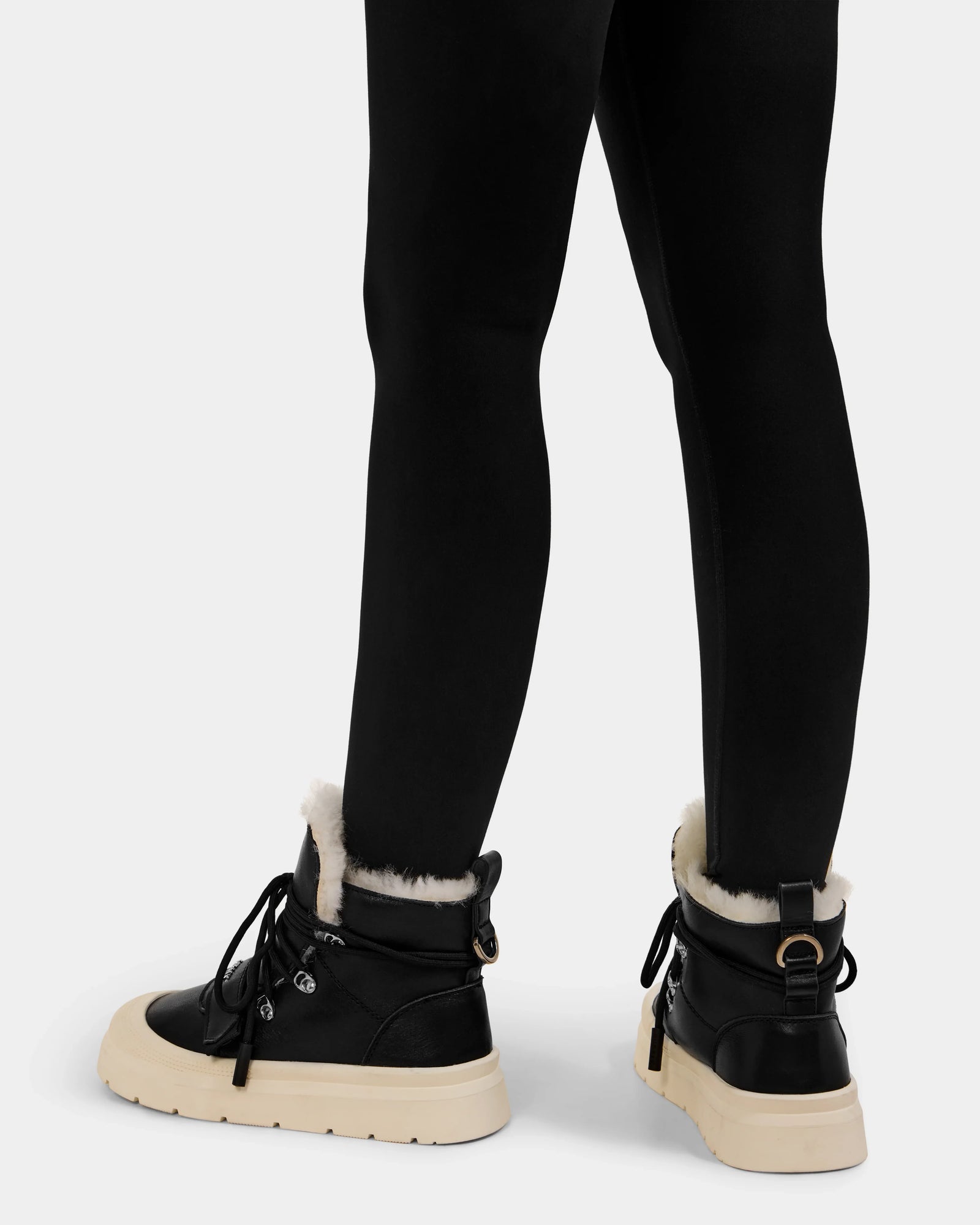 A person wearing black leggings and Rudsak’s ALEXEY L - L225518 Black women’s winter boots with white wool lining and beige chunky soles, shown from the back against a plain light background.