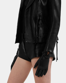 A person stands against a light background, wearing Rudsak’s ALEXIA - L425543 Black leather jacket and sleek women’s gloves, styled with black shorts. Only the torso and upper legs are visible.