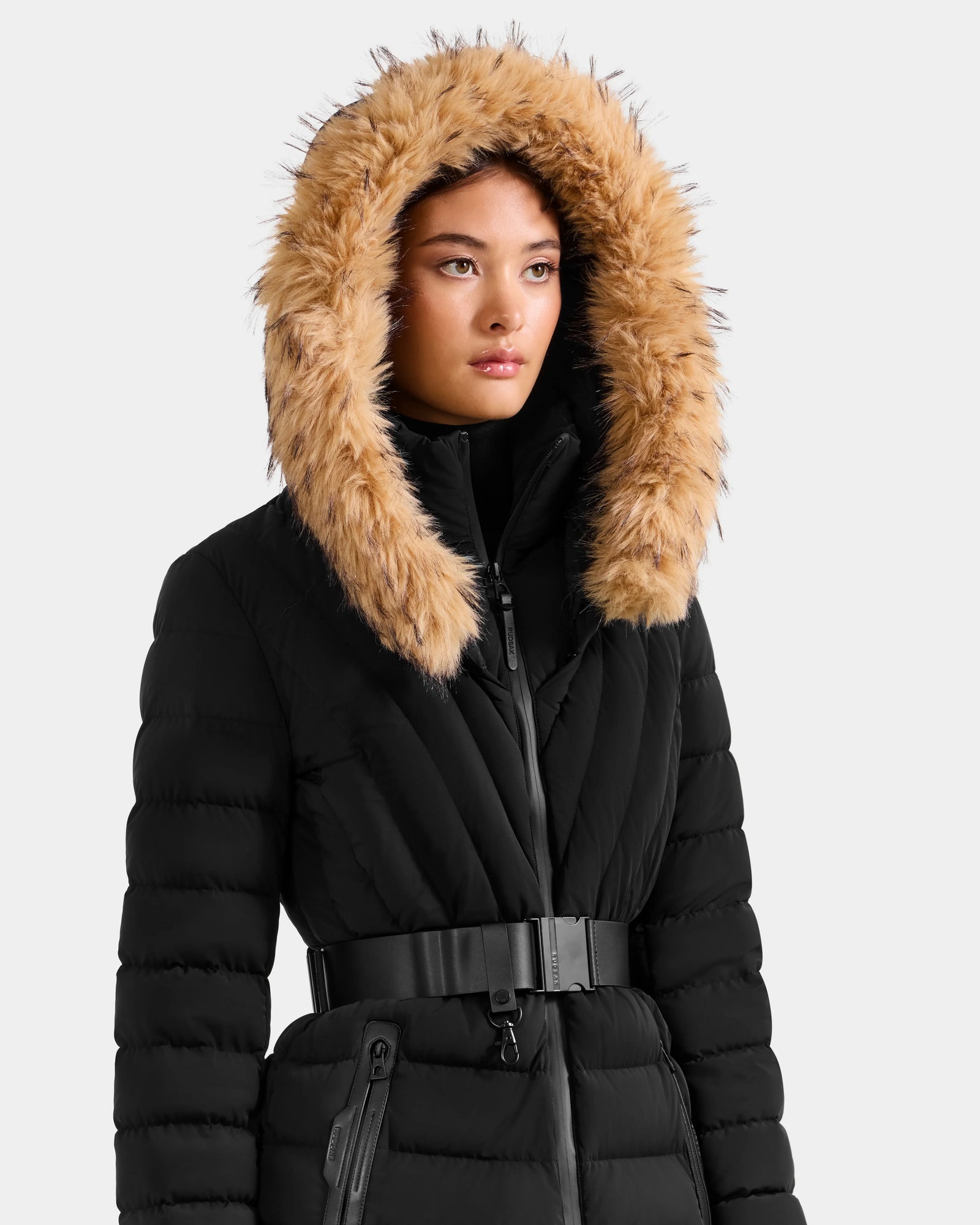 A woman wears the Rudsak ALISA F - L125527 Black belted quilted puffer coat with a large hood and tan faux-fur trim, standing against a plain light background.