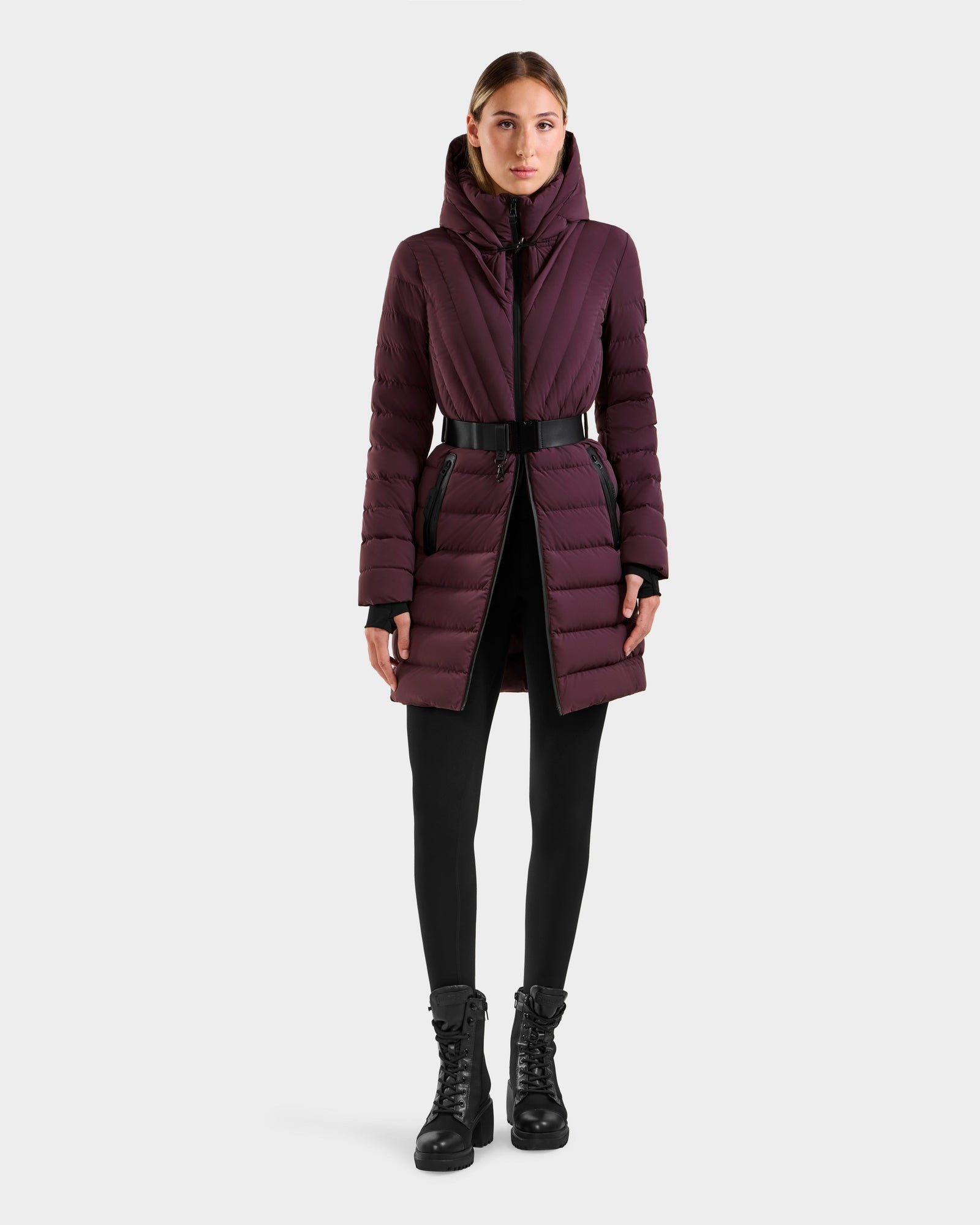 A woman in the Rudsak ALISA M - L125586 Bordeaux long belted down parka with black zipper details, black leggings, and black lace-up boots stands against a plain light background.