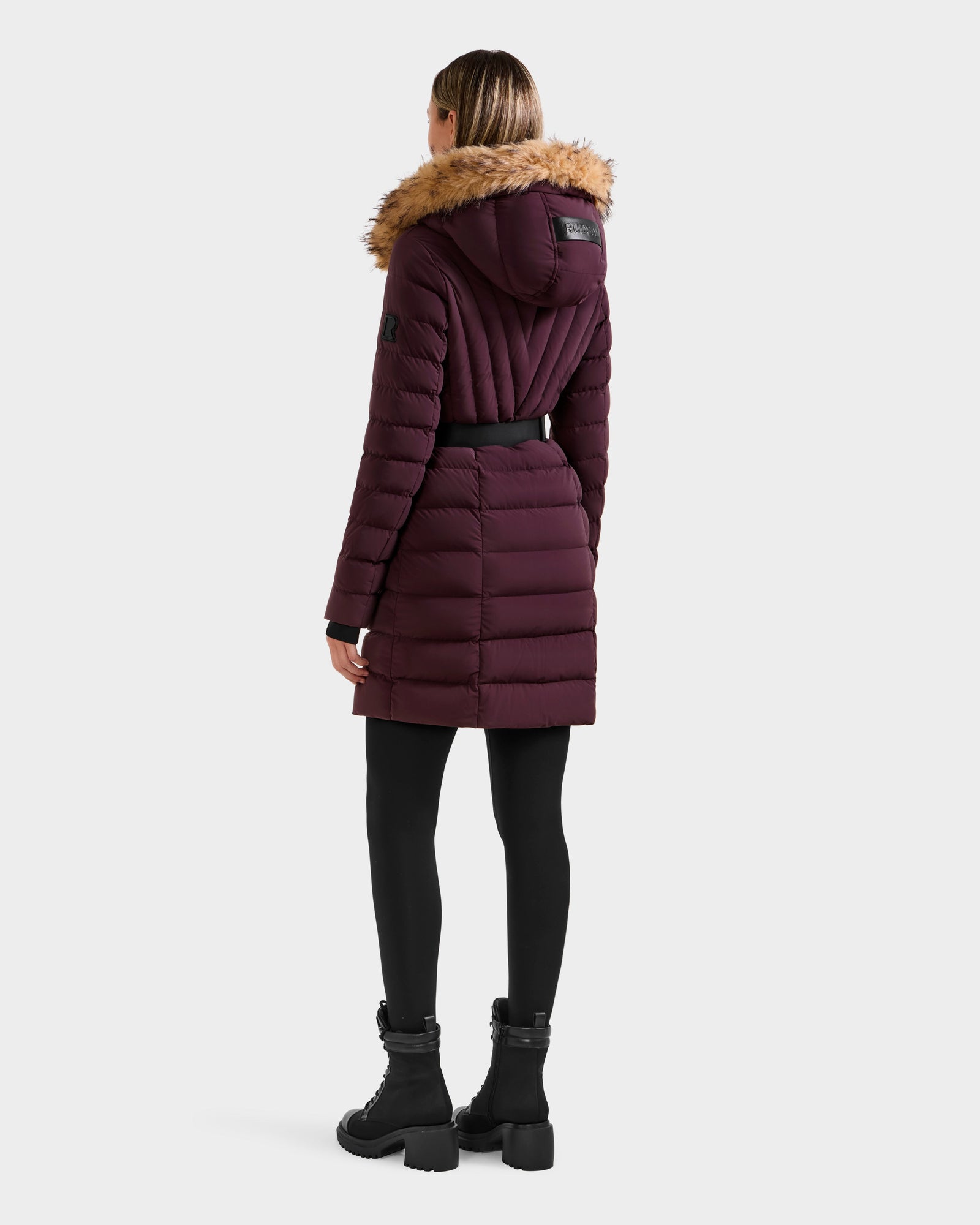 A woman wearing the Rudsak ALISA M - L125586 Bordeaux quilted down parka with faux fur-trimmed hood and vegan leather belt stands facing away from the camera, paired with black leggings and boots against a plain background.