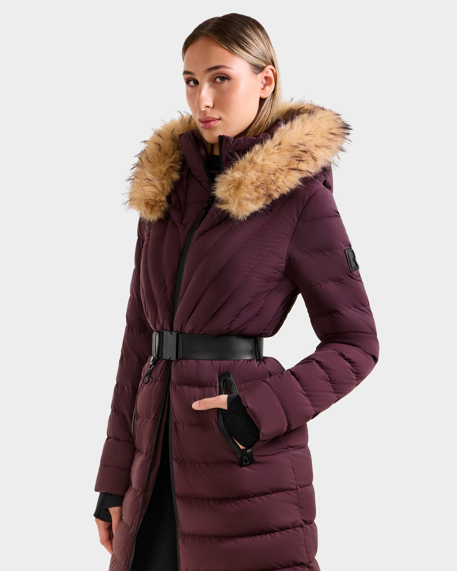 A woman wears the Rudsak ALISA M - L125586 Bordeaux, a maroon quilted down parka with a vegan leather belt and large faux fur-trimmed hood, standing against a plain light background.