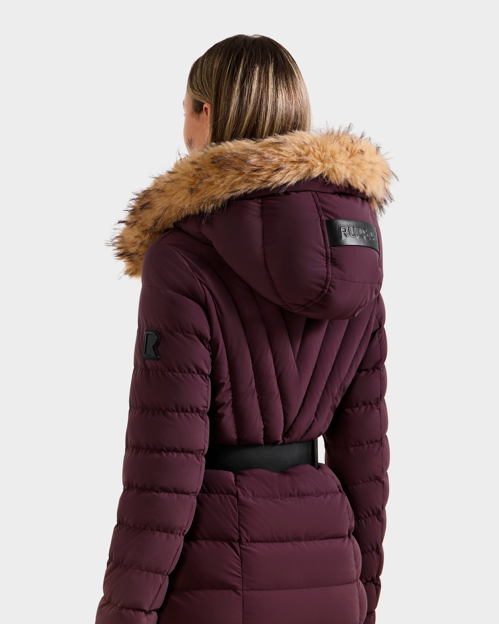 A person is seen from the back wearing the Rudsak ALISA M - L125586 Bordeaux parka, featuring a maroon quilted design with a faux fur hood, fitted waist with vegan leather belt, and visible stitching for a cozy winter look.