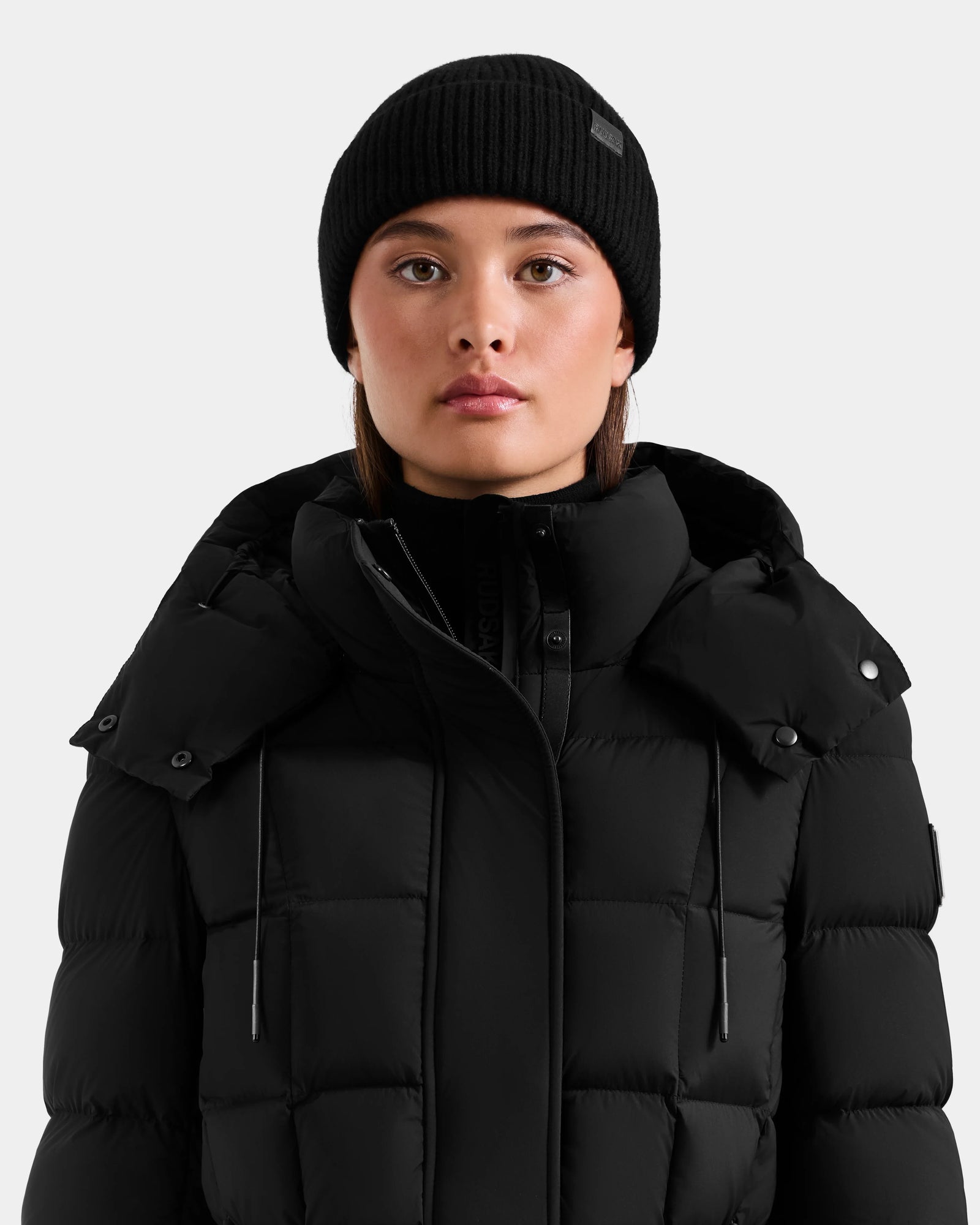 A person faces forward against a plain light gray background, wearing the Rudsak ANDREY - U425507 Black puffer jacket and a black unisex beanie.