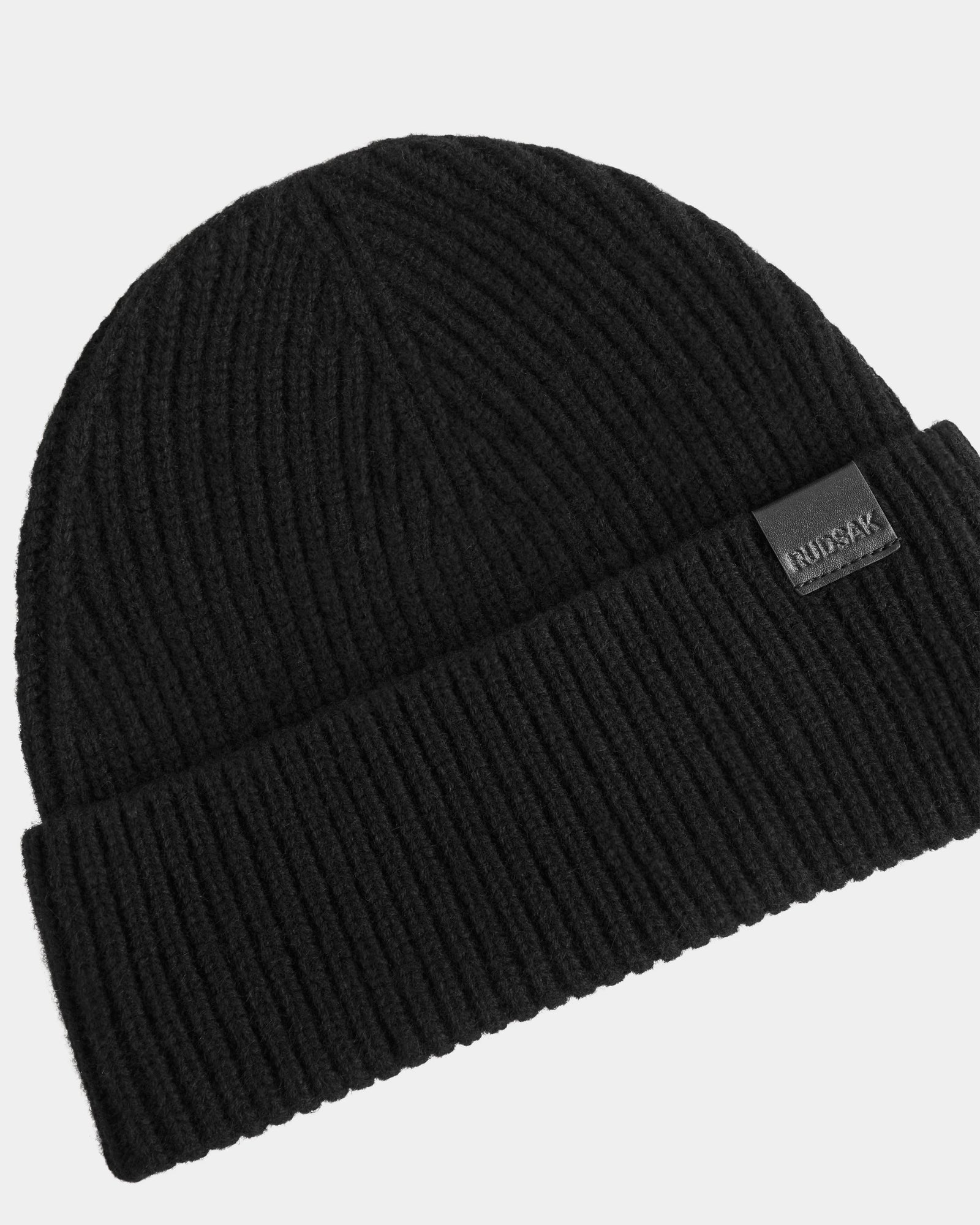The ANDREY - U425507 Black by Rudsak is a unisex black ribbed knit beanie with a folded cuff, featuring a small rectangular Rudsak logo patch on the cuff for classic style and comfort.