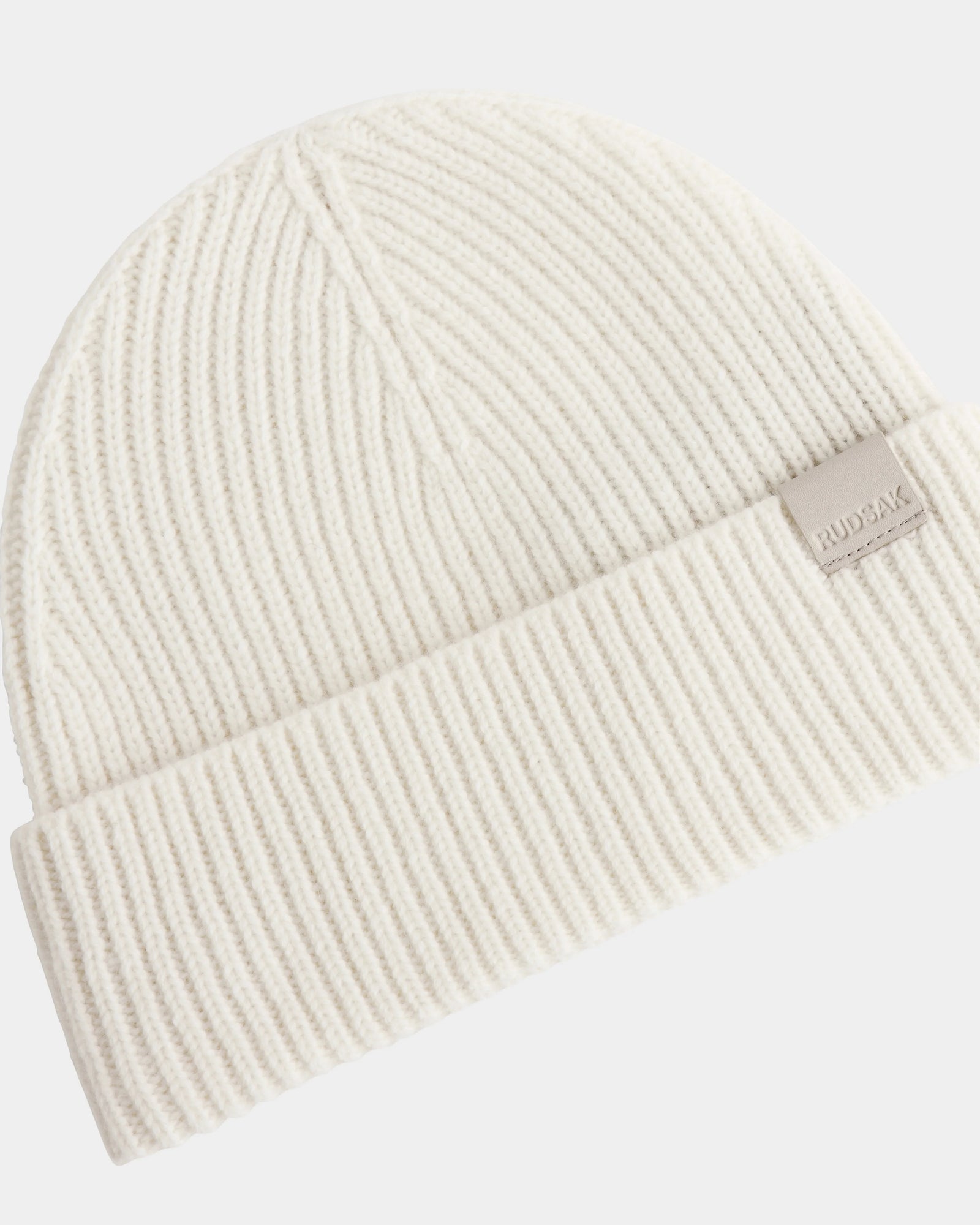 The ANDREY - U425507 Vanilla by Rudsak is a cream ribbed knit unisex beanie with a folded brim and a small rectangular RUDSAK tag on the cuff.