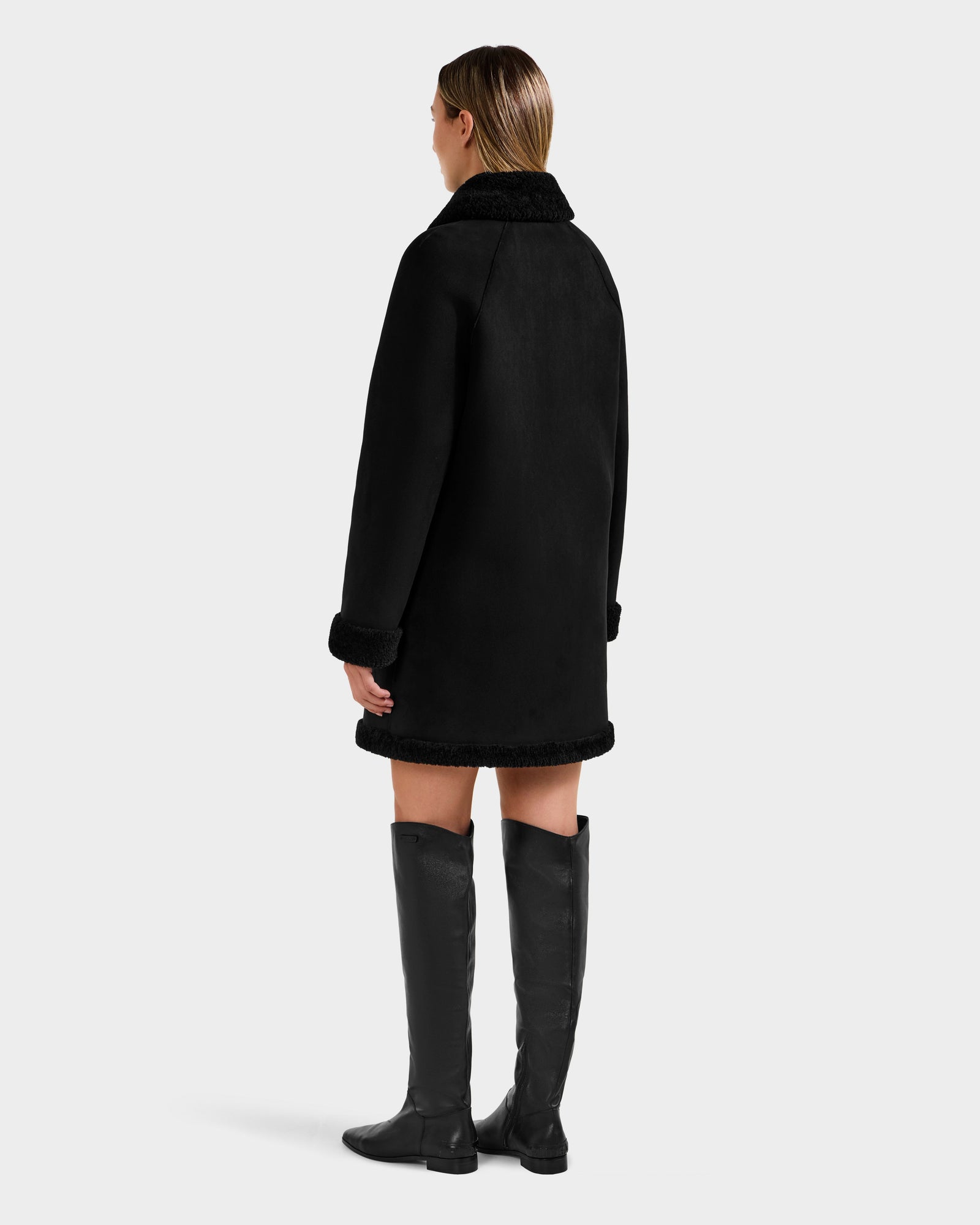 A woman in the Rudsak ANGILICA - L125600 Black reversible knee-length coat with faux shearling collar and cuffs and knee-high black leather boots stands facing away, her straight, shoulder-length hair against a plain light gray background.