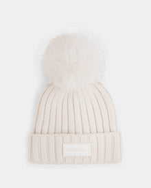 The ANISA - L423500 Vanilla by Rudsak is a cream-colored ribbed knit beanie with a plush faux fur pom pom and folded brim, accented by a rectangular Rudsak label—perfect for elevating your winter wardrobe.
