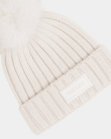 The ANISA - L423500 Vanilla by Rudsak is a cream ribbed knit beanie featuring a large faux fur pom pom and a folded brim with a rectangular Rudsak logo patch—an essential addition to your winter wardrobe.
