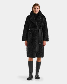 A woman faces forward in the Rudsak ANNABELLE FSU - L125512 Black teddy trench coat, black turtleneck, and black knee-high boots against a plain light gray background.